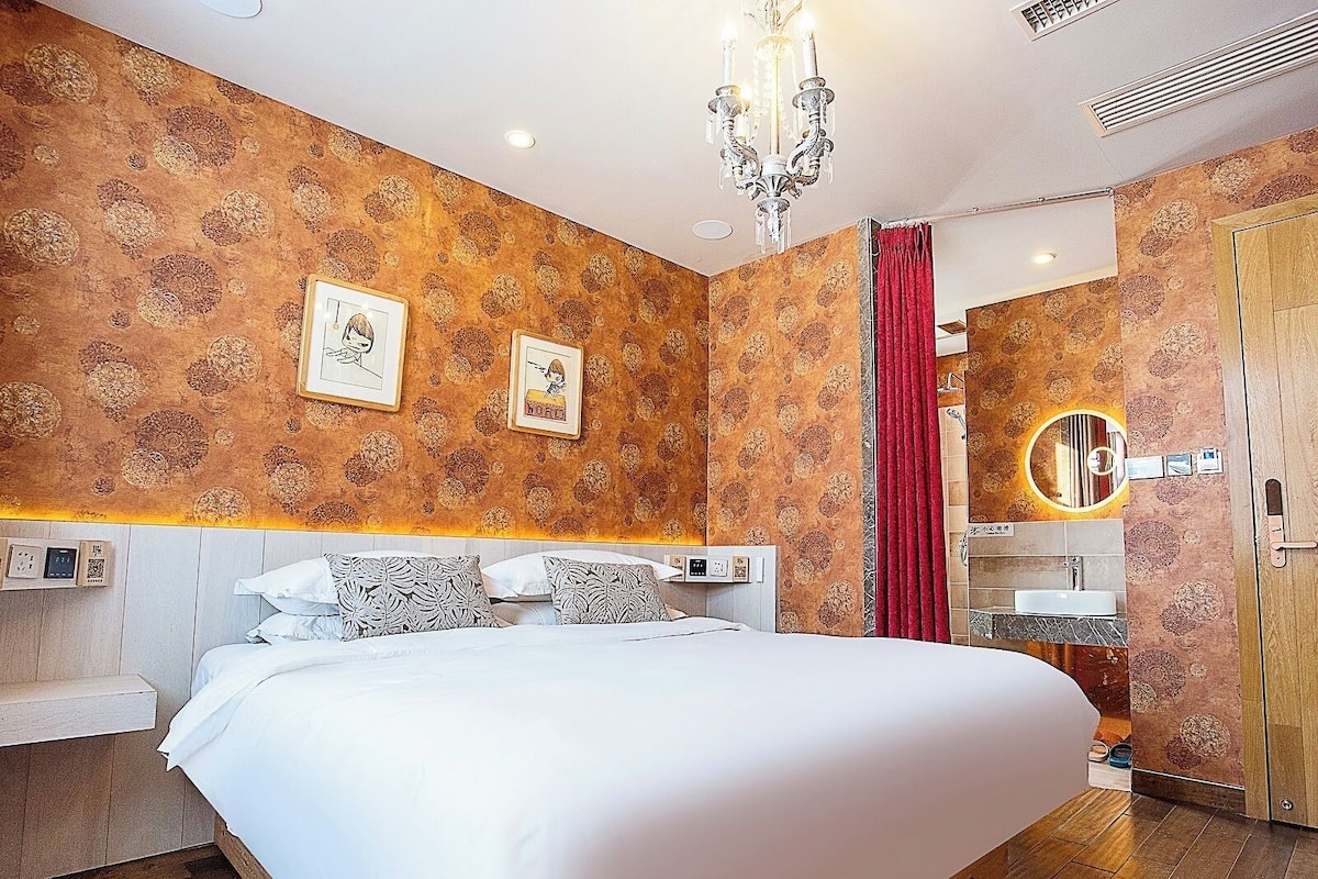 A bedroom with a large bed, a chandelier, a red curtain, and a bathroom with a sink and mirror.