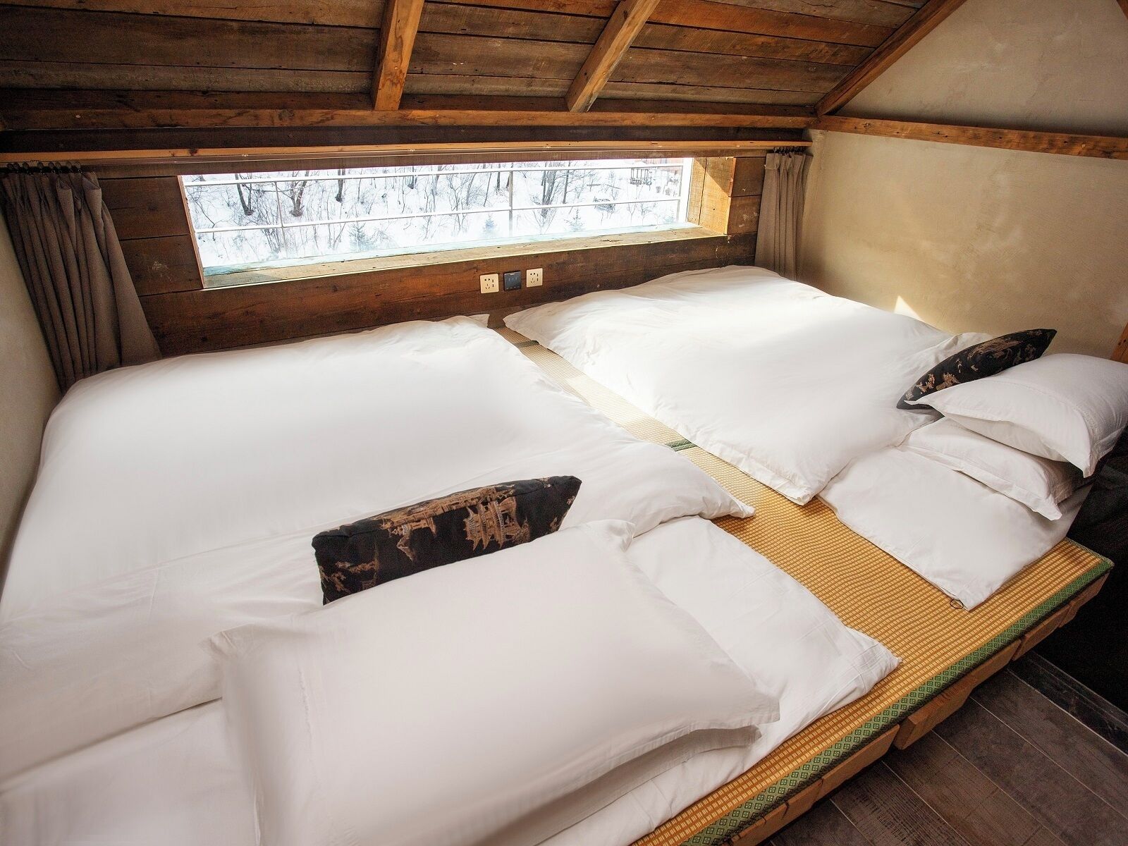 A double bed with white linens and two decorative pillows, a window with a view of trees, and wooden beams on the ceiling.