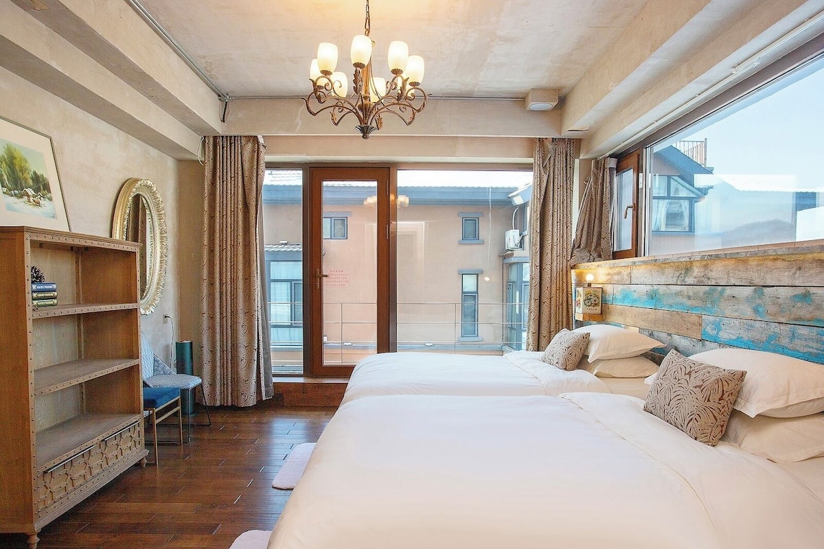 A bedroom with a large bed, wooden headboard, a chandelier, a bookshelf, and a view of buildings through the window.