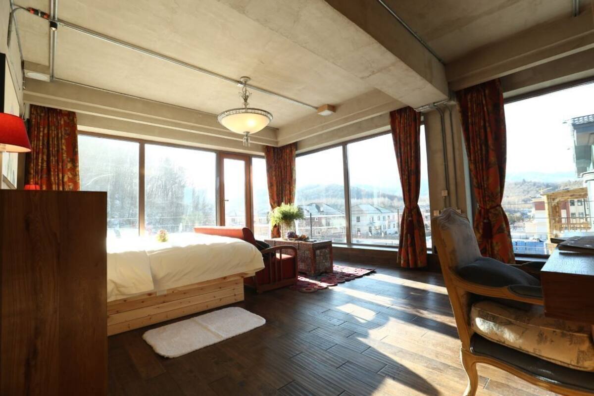 A spacious bedroom with a large bed, a wooden desk, a chair, and a view of the city through large windows.