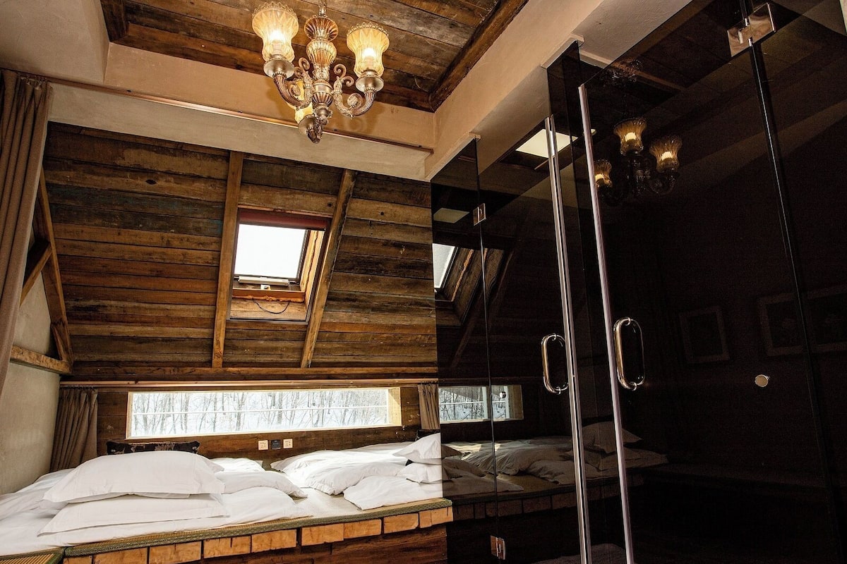 A bedroom with a wooden ceiling, a chandelier, a large window, a glass shower, and a bunk bed.