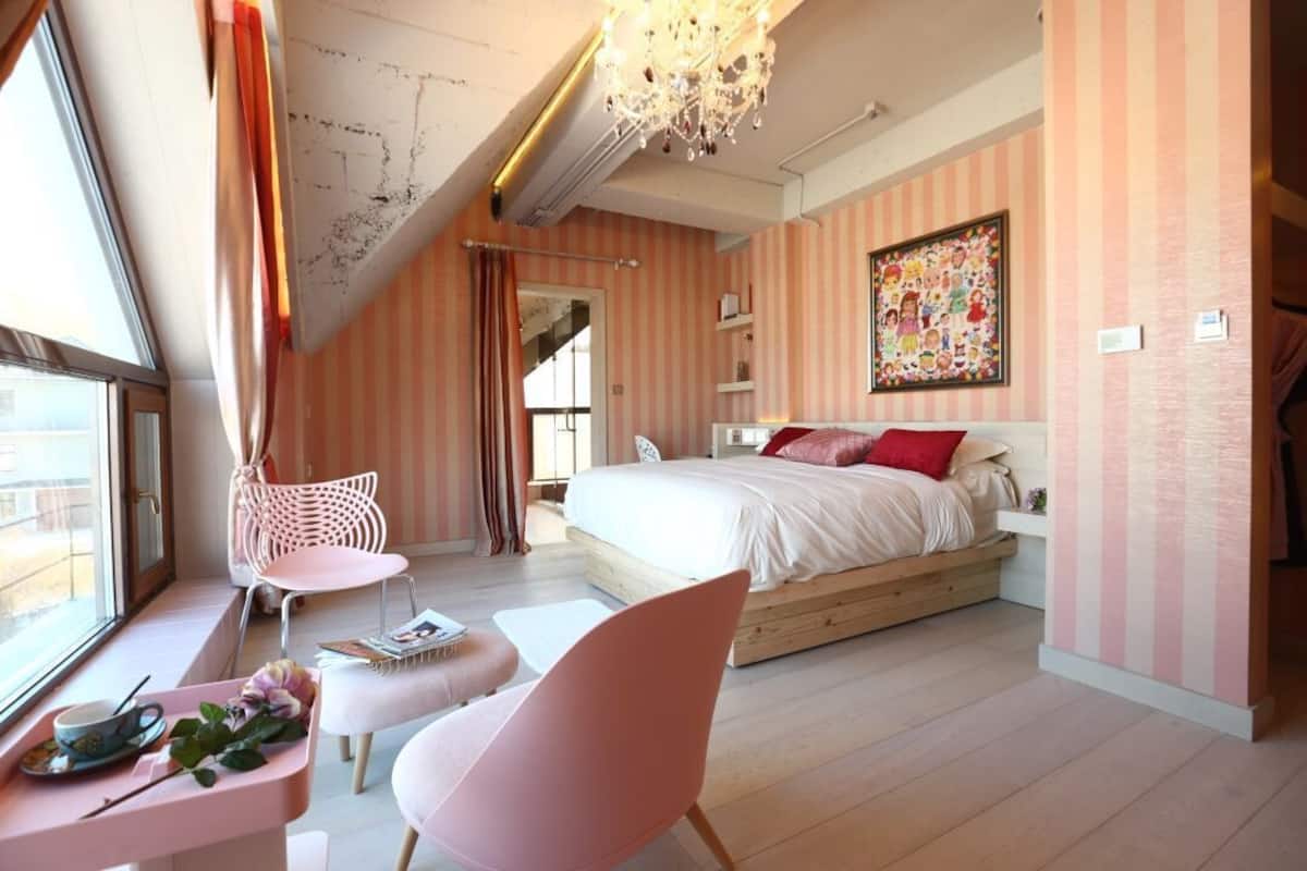 A bedroom with a large bed, a window, a pink chair, a wooden floor, and a chandelier.