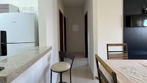 Un couloir étroit mène à une cuisine équipée d’un réfrigérateur, à un coin repas avec une table et des chaises, et à un meuble de couleur sombre.
