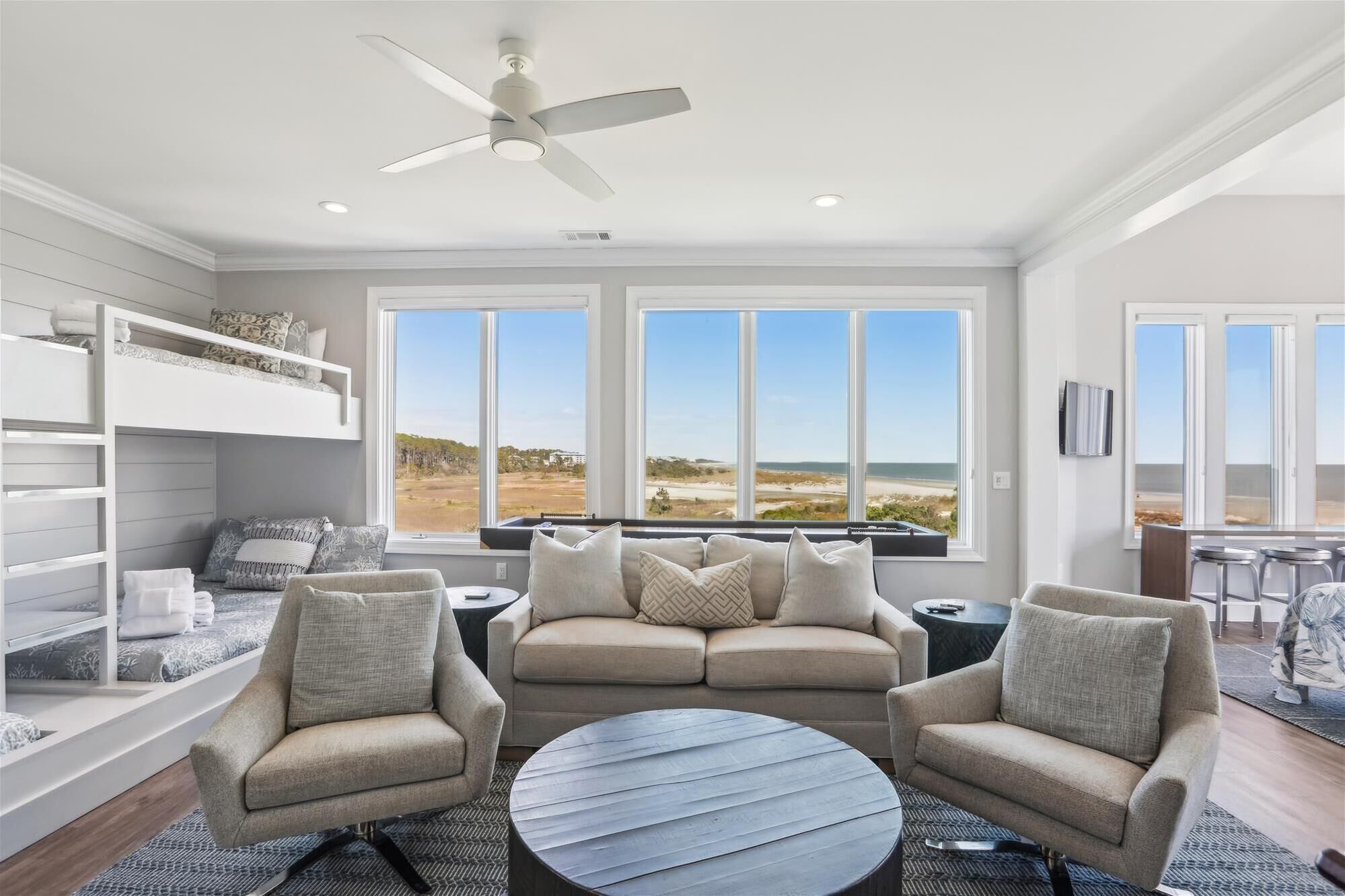 Un salon moderne avec un canapé, deux fauteuils, une table basse et un ventilateur de plafond. De grandes fenêtres offrent une vue sur la plage.