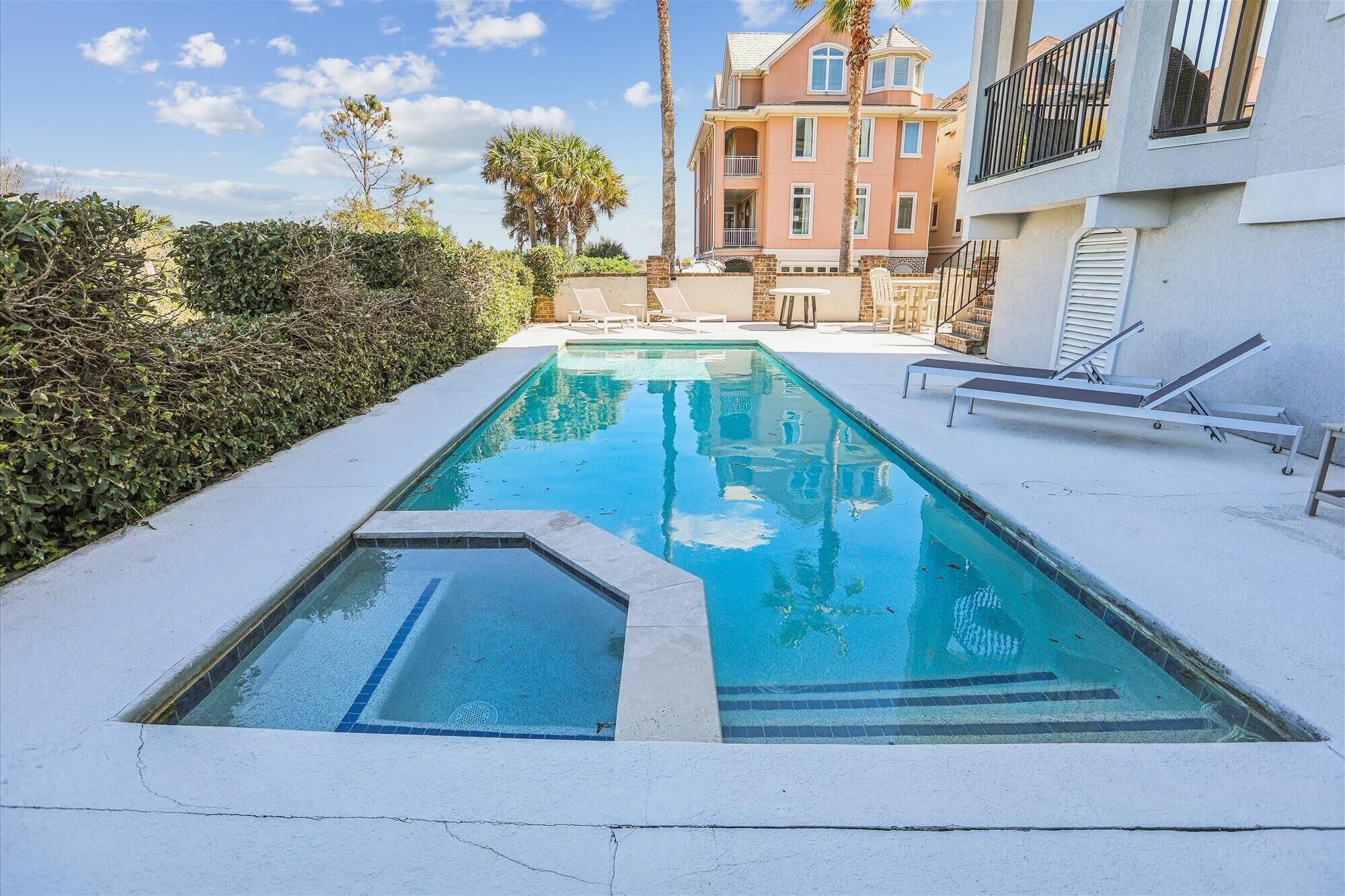A rectangular swimming pool with a tiled edge, surrounded by a white deck. There are lounge chairs and a small table on the deck. In the background, there is a multi-story building with balconies and a palm tree.