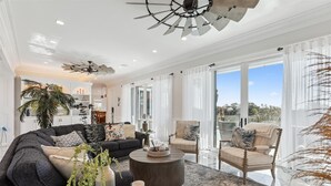 A spacious living room with a dark grey sofa, a round coffee table, and a large window offering a view of the outdoors. - 8 Sea Front, Oceanfront Estate, Pool, Huge Game Room, Elevator (Hilton Head Island)