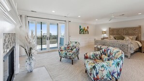 A spacious bedroom with a large bed, a sitting area with floral-patterned chairs, a fireplace, and a balcony with a view. - 8 Sea Front, Oceanfront Estate, Pool, Huge Game Room, Elevator (Hilton Head Island)