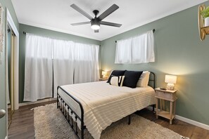 Un dormitorio con una cama, dos mesitas de noche con lámparas, un ventilador de techo y una ventana con cortinas.