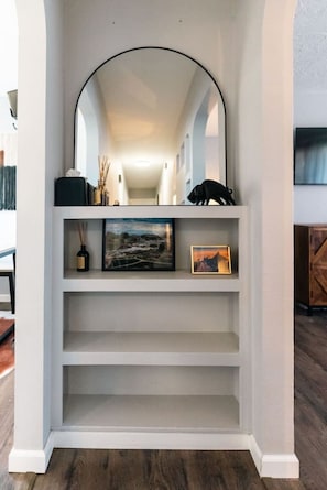 A white shelving unit with a mirror above it, a picture frame, and a small black object on the top shelf. - Sioux Falls Midtown Escape (Sioux Falls)