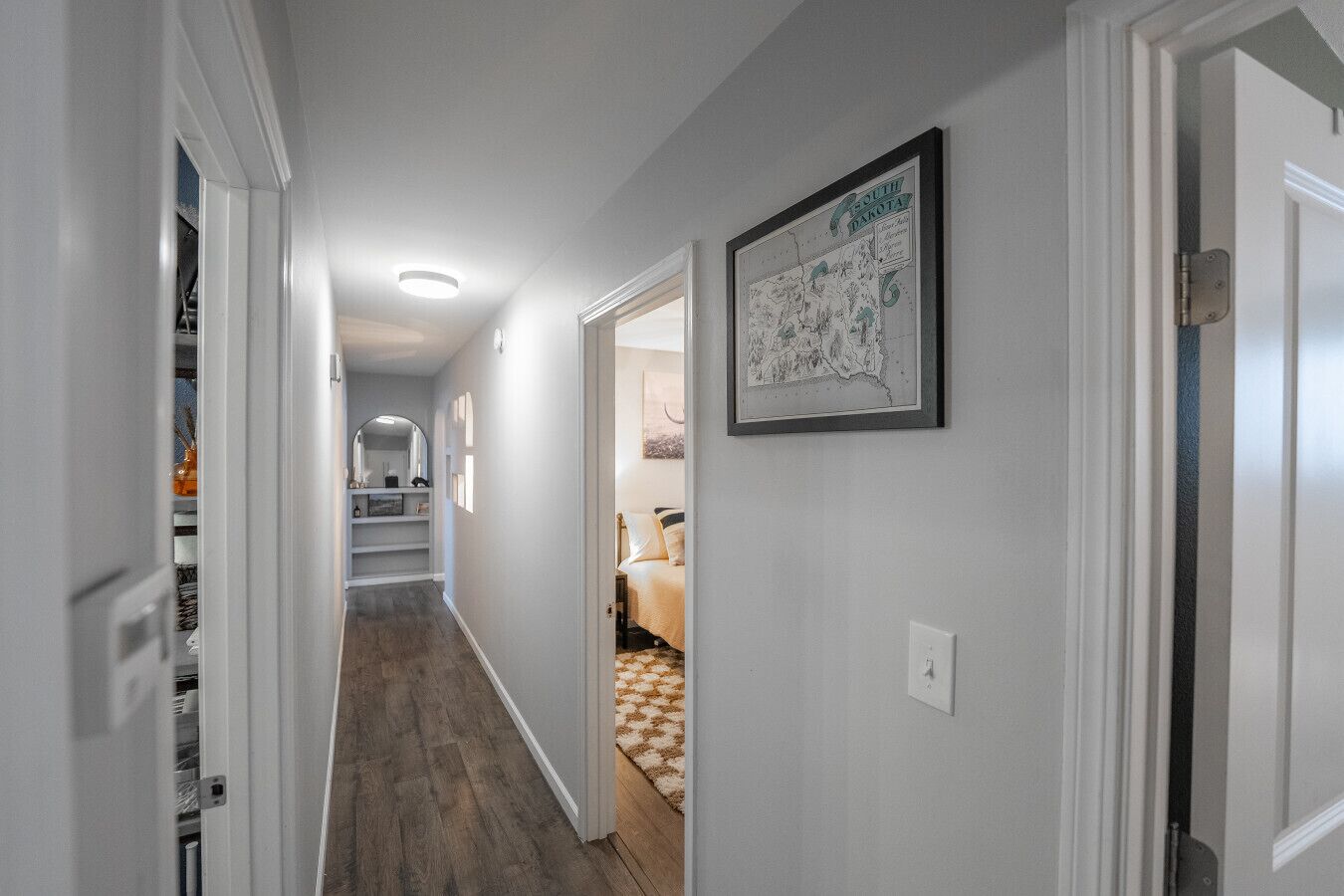 A hallway with a wooden floor, a framed map on the wall, and a door leading to a bedroom with a bed and a nightstand.