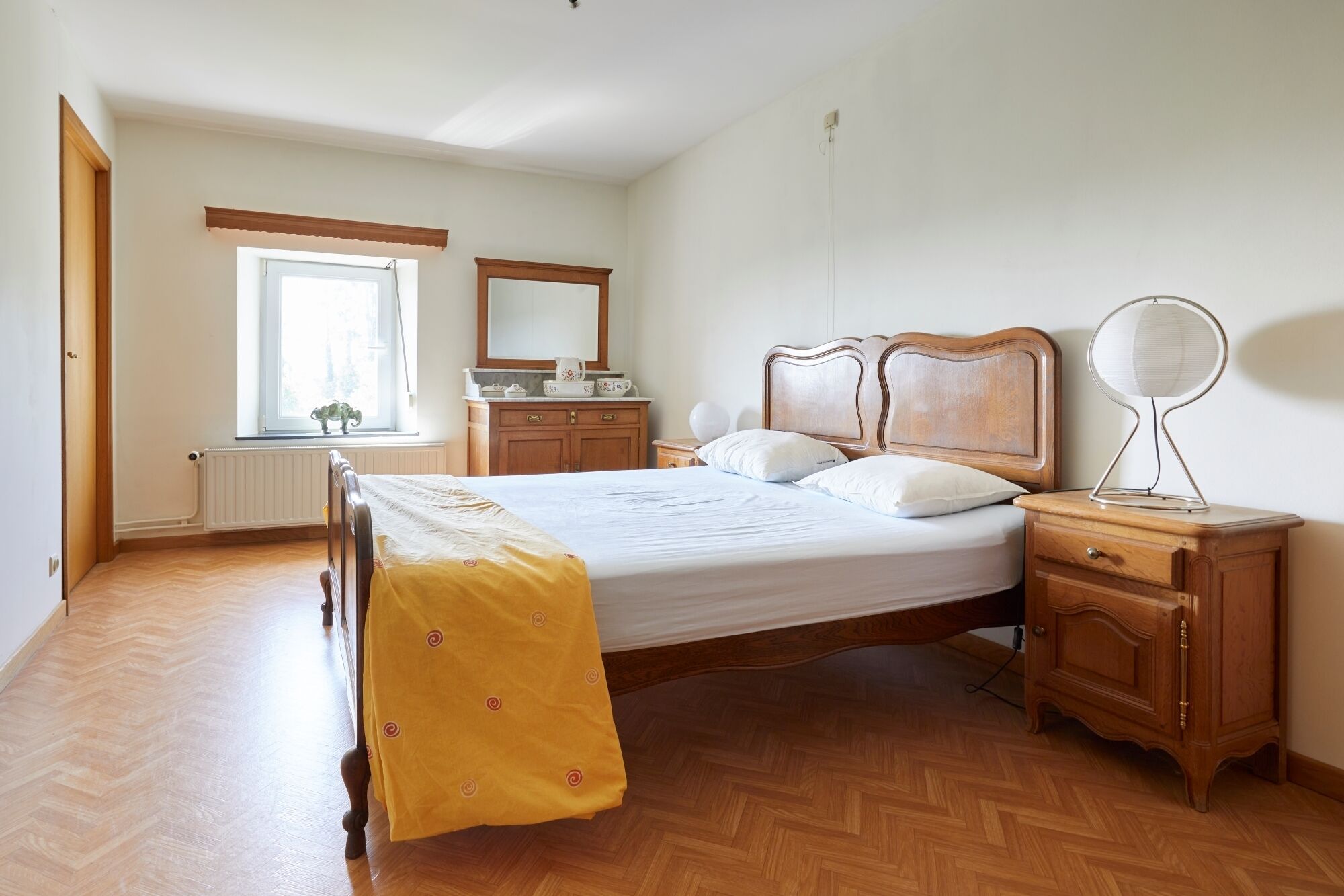 Ein Schlafzimmer mit einem Holzbett, einem Nachttisch mit Spiegel, einer Kommode und einem Fenster mit Blick ins Freie.