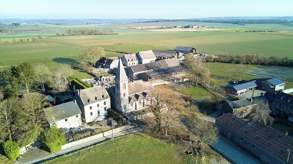 An aerial view of a historic estate with multiple buildings, including a prominent tower, surrounded by expansive green fields and a clear sky.