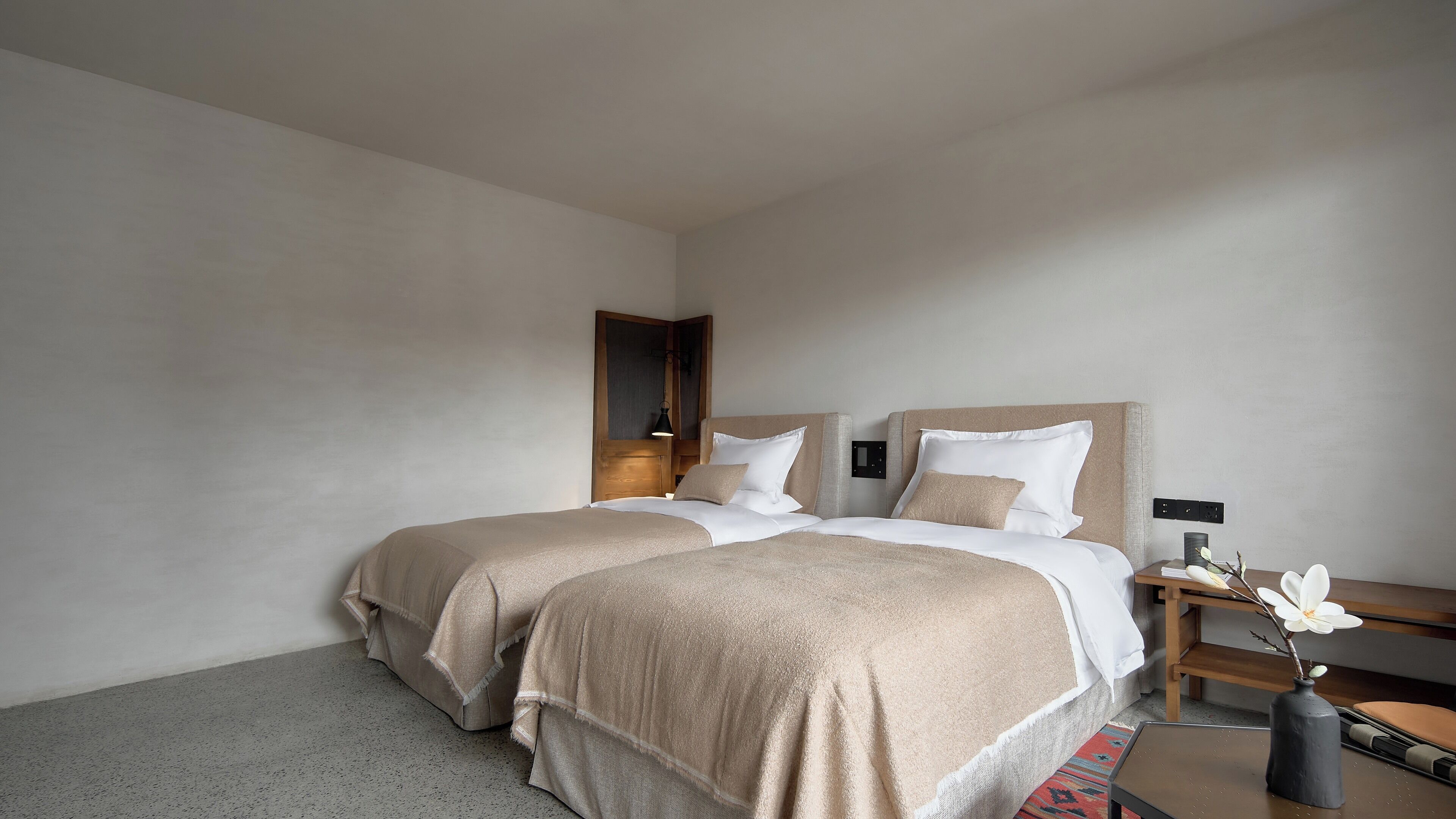 A hotel room with two beds, a nightstand, a lamp, and a vase with flowers.