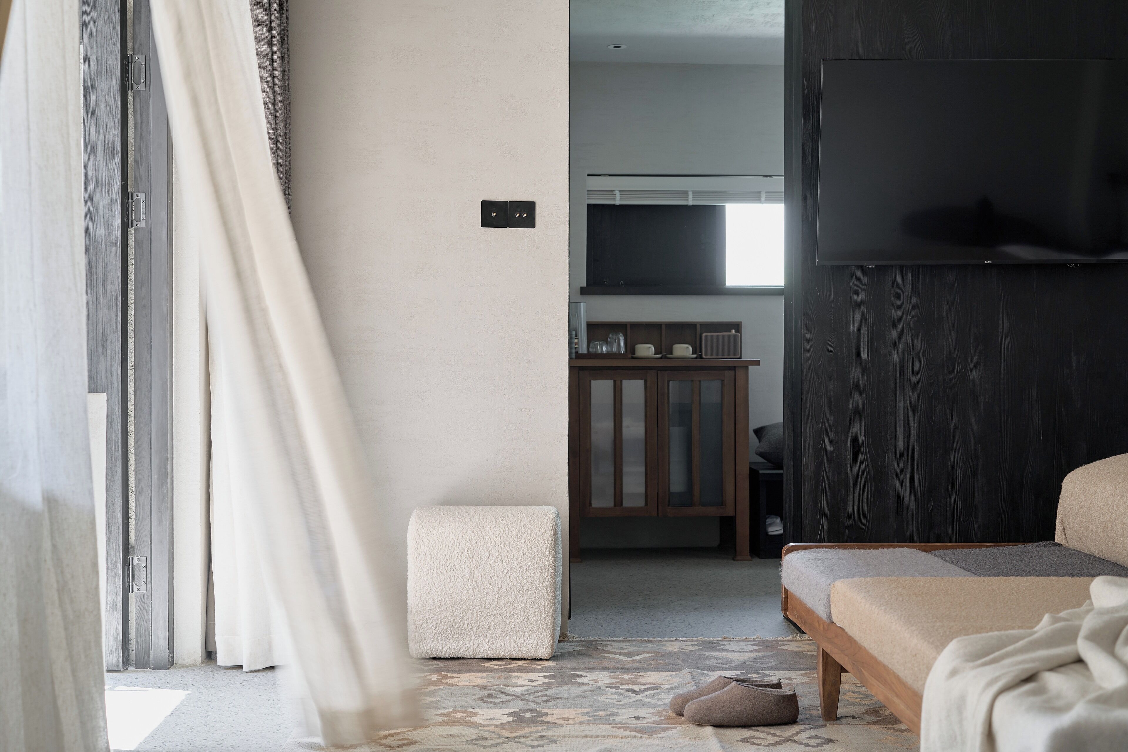 A modern hotel room with a bed, a small seating area, a television, and a view of the hallway.