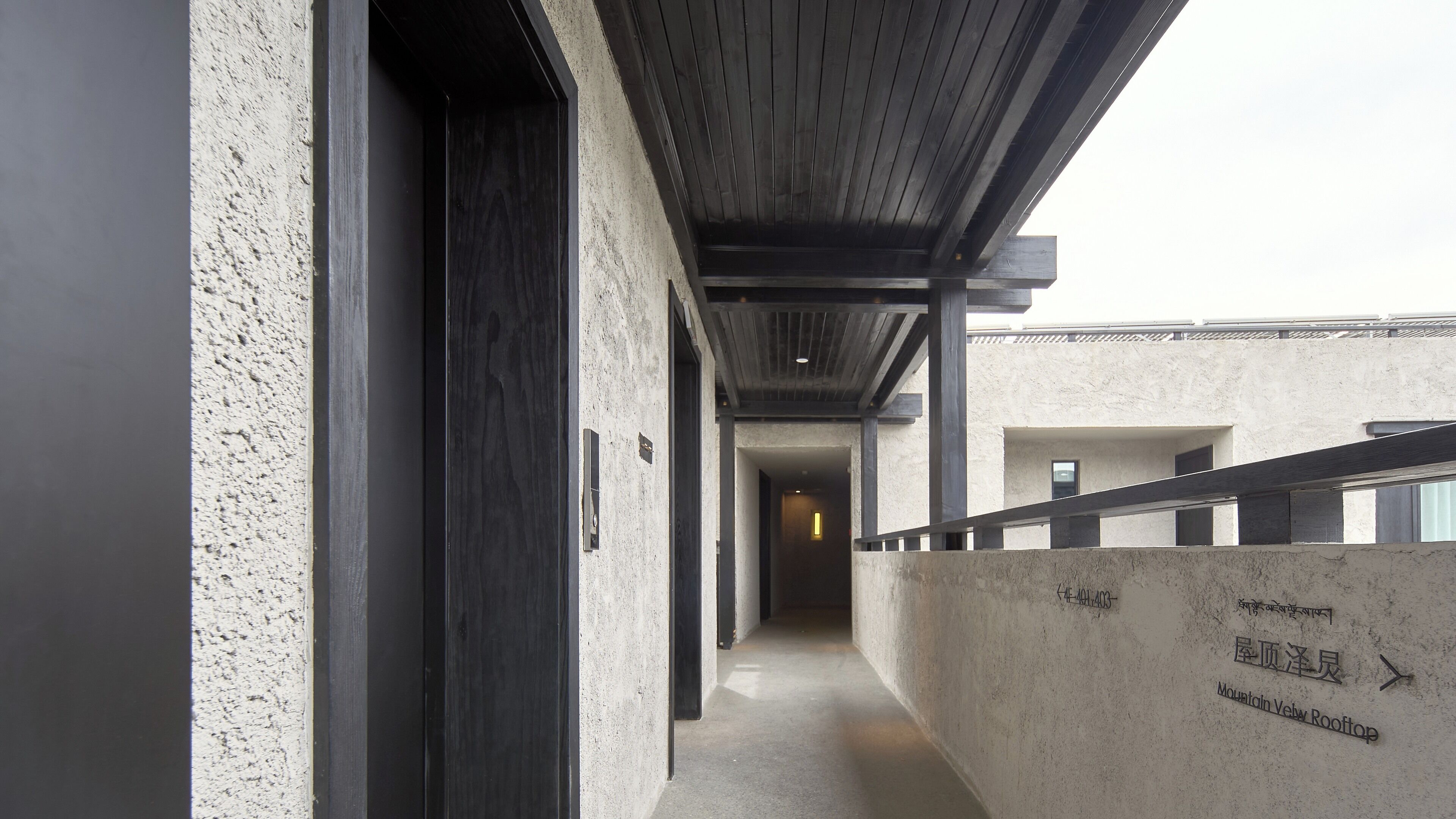 Un pasillo con pared de concreto y techo de madera, que lleva a una puerta. Hay un cartel con texto y una flecha señalando a la derecha.