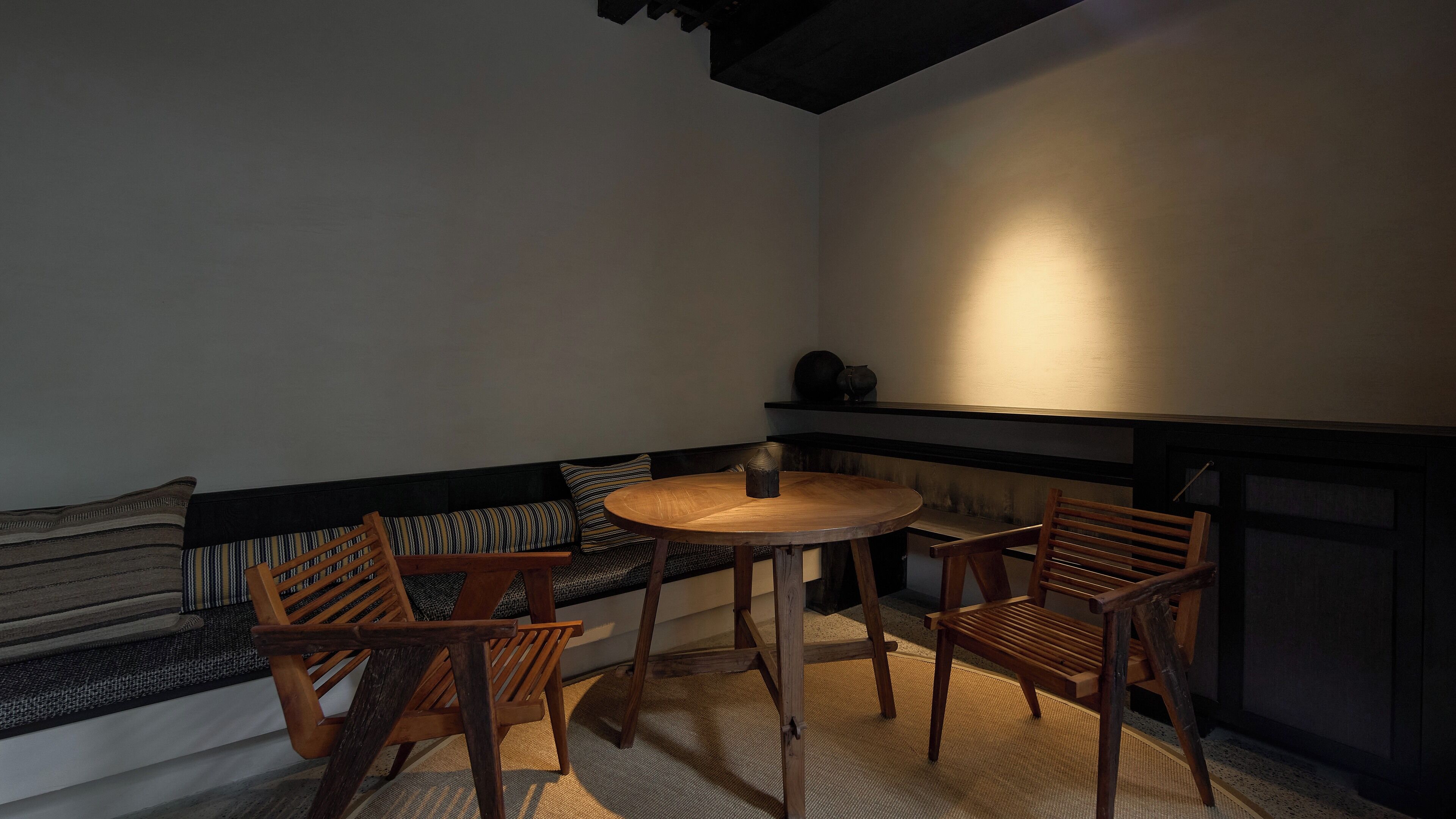 Un coin salon accueillant avec une table ronde en bois et deux chaises, un banc avec des coussins à rayures et un mur foncé sur lequel est fixée une étagère.