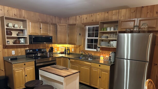 A kitchen with wooden cabinets, a stainless steel refrigerator, a microwave, and a stove. There are two stools and a small island with a cutting board.