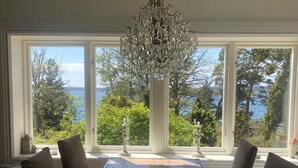 A dining area with a wooden table, chairs, and a chandelier. Large windows offer a view of trees and a body of water. - Stockholm  Archipelago – Idyllic Island (Värmdö)