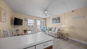 A beach house interior with a dining area, a sofa, a TV, and a ceiling fan. - Emerald Skye 43 | Gulf-Front + Pool (Orange Beach)
