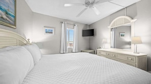 Une chambre d’hôtel équipée d’un lit, d’une commode, d’un miroir, d’un ventilateur de plafond, d’une télévision et d’une fenêtre avec des rideaux.