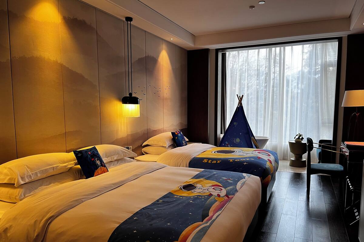 Une chambre d’hôtel avec deux lits, une table de chevet, une lampe, une fenêtre avec des rideaux et une petite table sur laquelle est posée une plante.