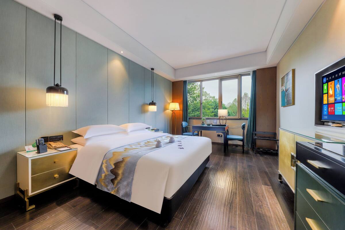 Une chambre d’hôtel avec un grand lit, des tables de chevet, un bureau, une chaise, une télévision et une fenêtre donnant sur des arbres.