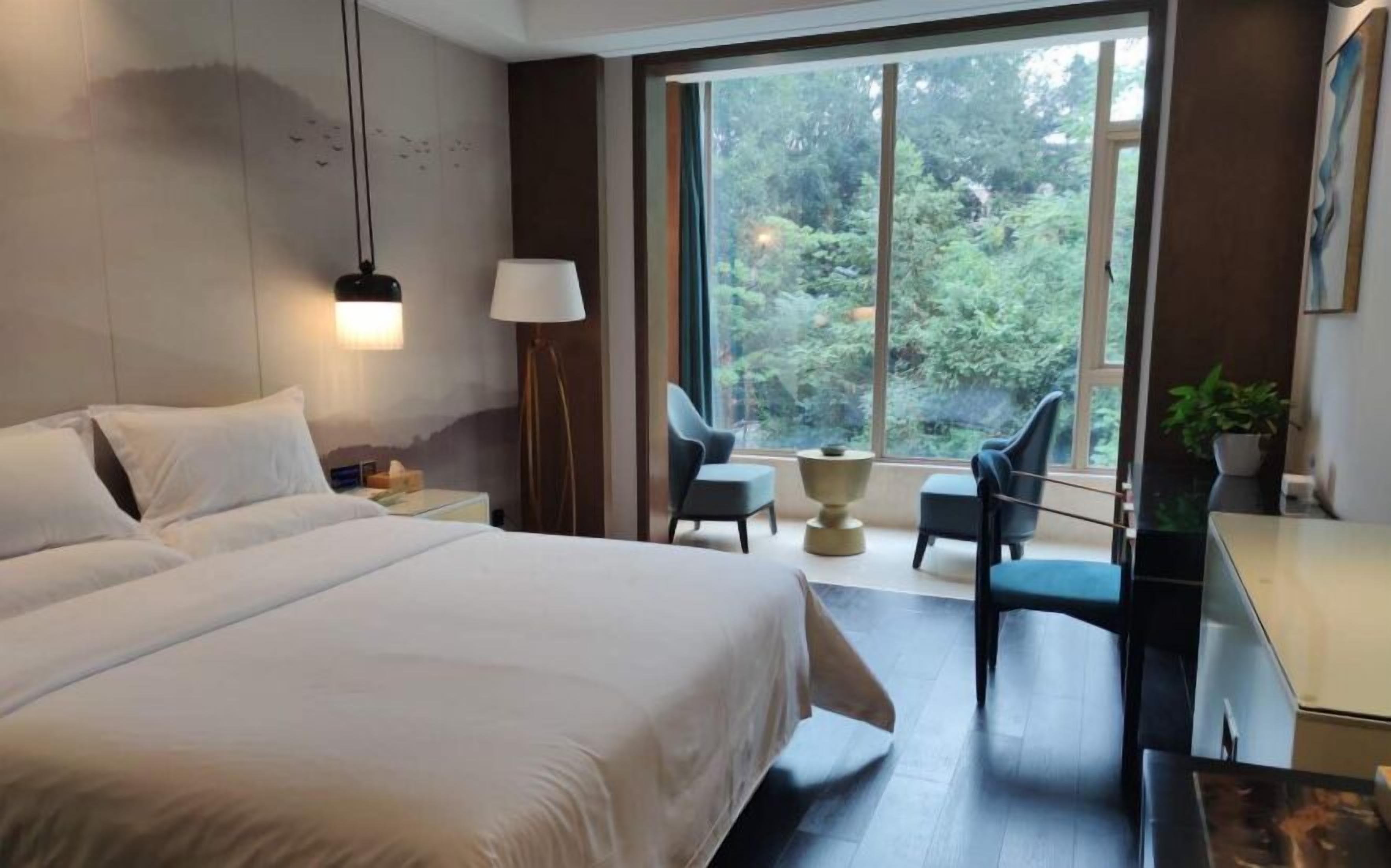 Habitación de hotel con una cama grande, dos sillones, un escritorio y vistas a zonas verdes por la ventana.