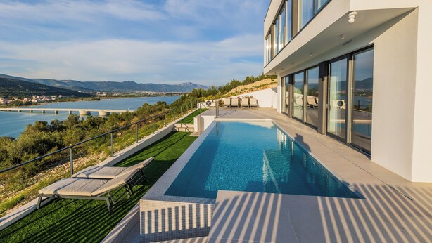 A modern building with a swimming pool, overlooking a body of water and hills.