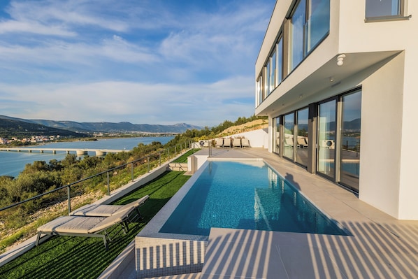 A modern building with a swimming pool, overlooking a body of water and hills.
