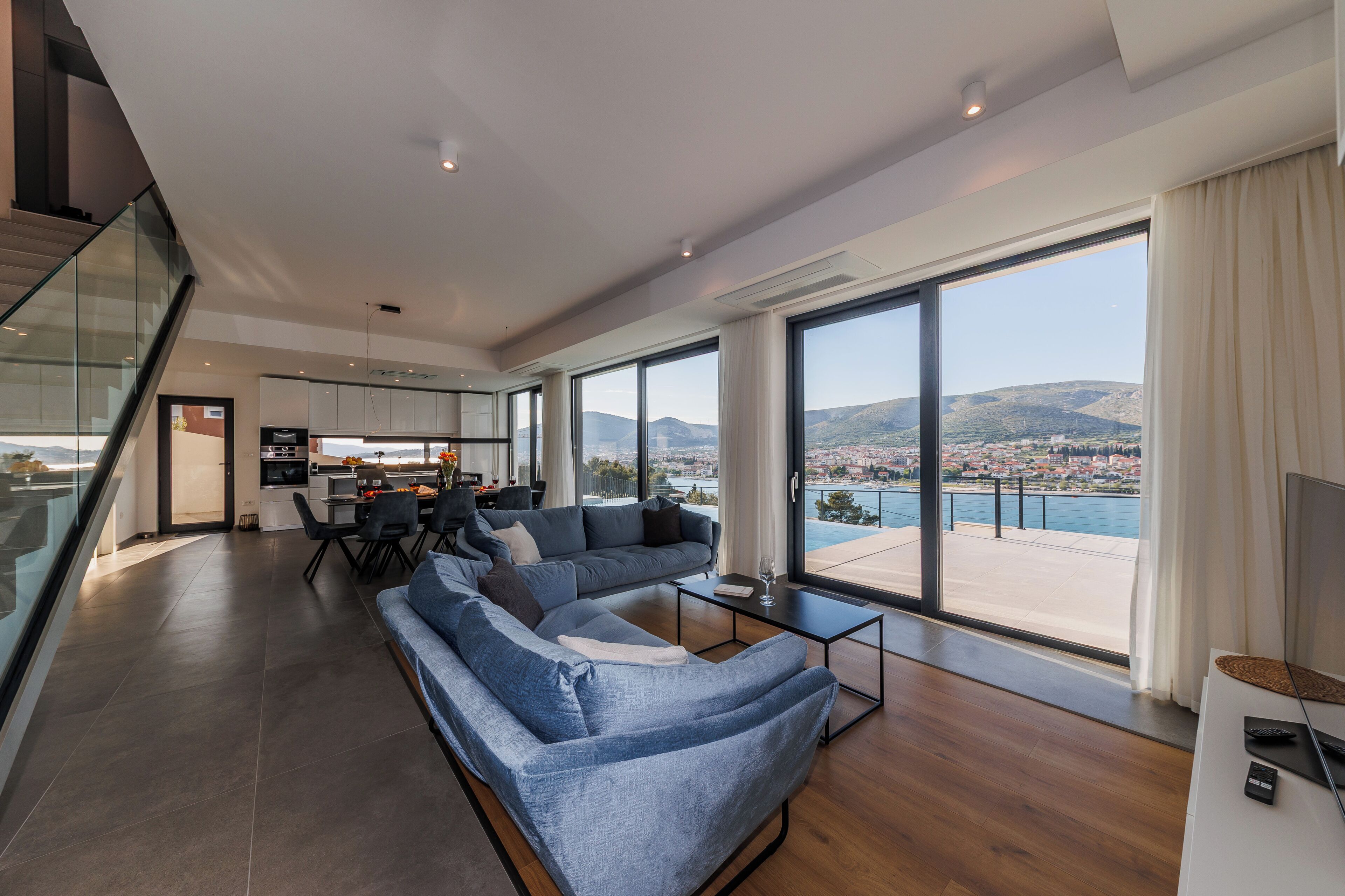 Ein modernes Wohnzimmer mit großem Sofa, Essbereich und Küche. Ein großes Fenster bietet Blick auf ein Gewässer und Hügel.