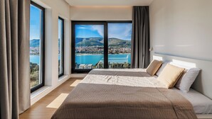 A bedroom with a large bed, a view of a city and water, and large windows.