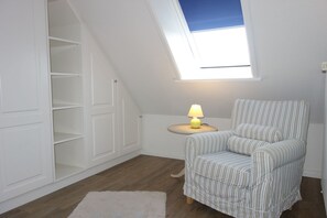 Ein Zimmer mit schrägen Wänden, einem Dachfenster, einem gestreiften Sessel, einem kleinen runden Tisch mit einer Lampe und eingelassenen weißen Regalen.