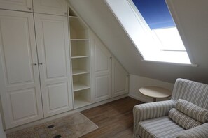 Ein Zimmer mit schräg angesetzter Decke, einem Oberlicht, einem eingebauten weißen Kleiderschrank, einem gestreiften Sofa und einem kleinen runden Tisch.