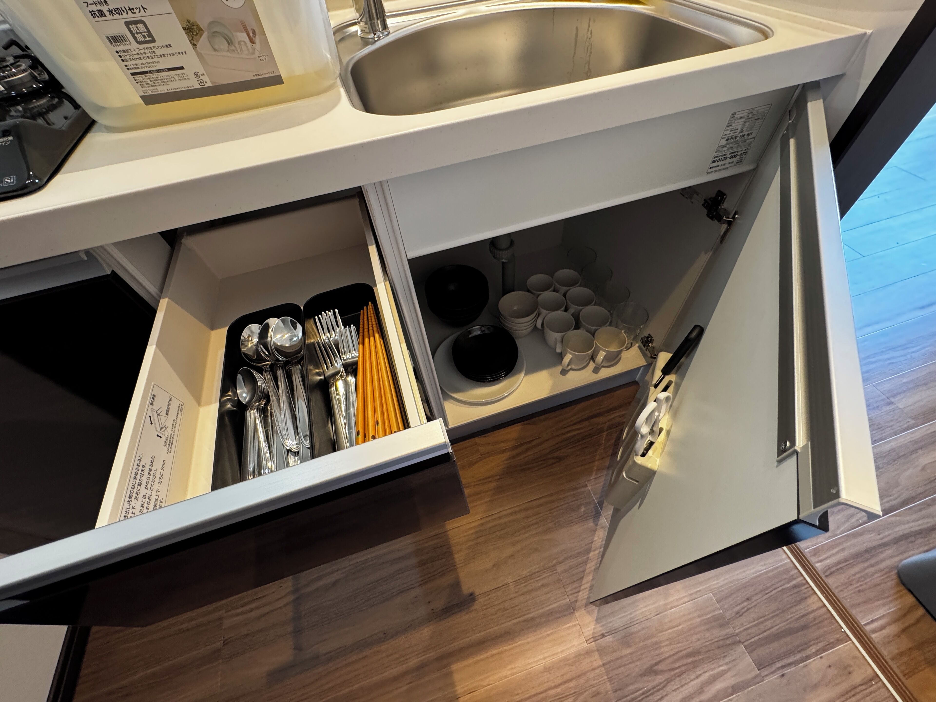 A kitchen with a stainless steel sink, a white countertop, and a drawer containing utensils and dishes.