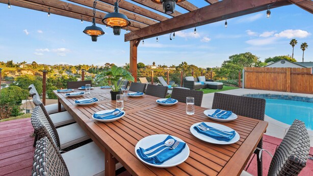 An outdoor dining area with a wooden table set for four, surrounded by wicker chairs, under a wooden pergola with hanging lights. - New! Spacious 5BD Home with Games, Pool & Hot Tub! (San Diego)