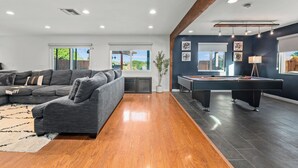 A modern living room with a sectional sofa, a pool table, and a large window offering a view of the outdoors. - New! Spacious 5BD Home with Games, Pool & Hot Tub! (San Diego)