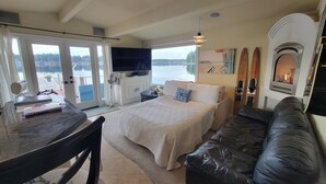 A bedroom with a bed, a black leather sofa, a dining table, and a TV. - Unique Lake Goodwin Waterfront Cabin with Private Hot Tub and Boat Moorage (Stanwood)