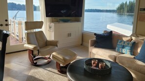 A living room with a large window overlooking a lake, a television, and a cozy seating area. - Unique Lake Goodwin Waterfront Cabin with Private Hot Tub and Boat Moorage (Stanwood)