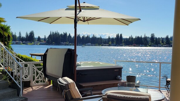 An outdoor deck with a table and chairs, a barbecue grill, and a large umbrella. - Unique Lake Goodwin Waterfront Cabin with Private Hot Tub and Boat Moorage (Stanwood)