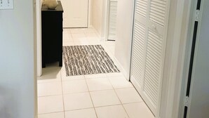 Un couloir doté d’un sol carrelé, d’un tapis noir et blanc, d’une porte blanche et d’une armoire blanche aux portes à persiennes.