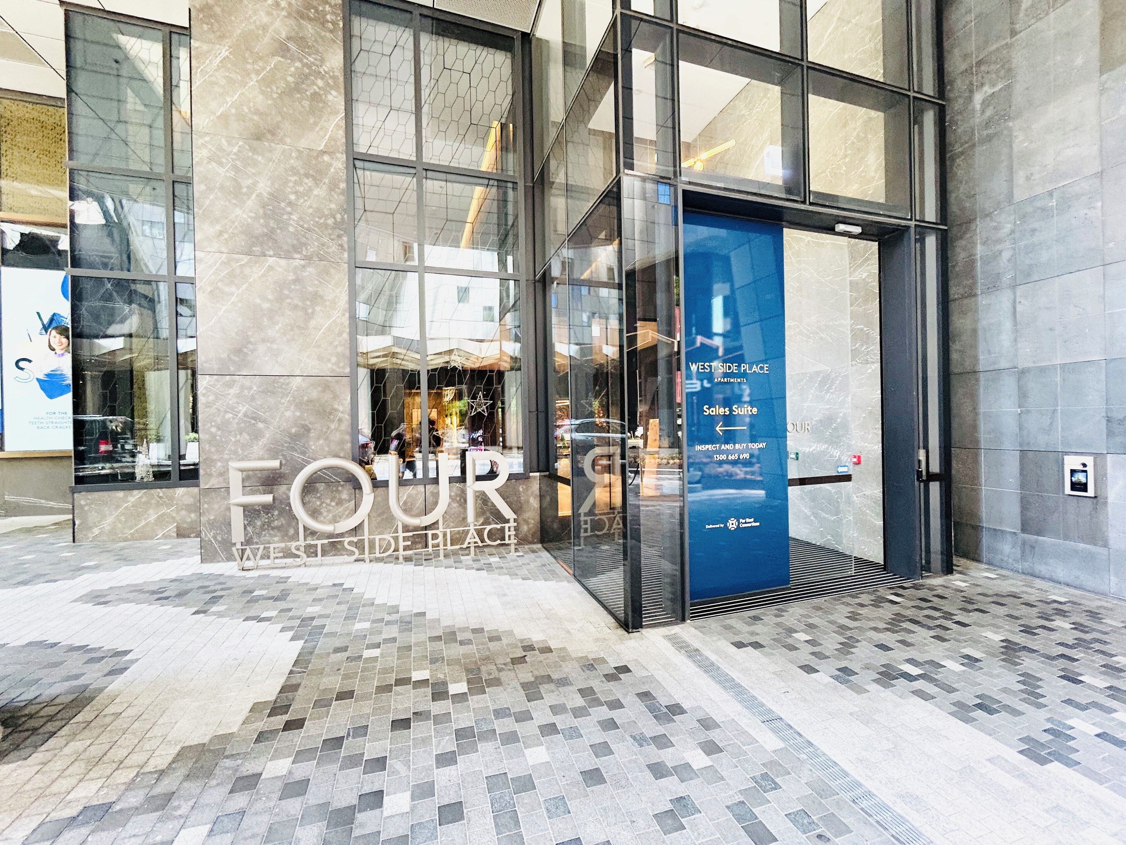 A modern building entrance with a glass door, a sign for 'West Side Place Apartments' and a 'Sales Suite' direction, and a tiled floor.
