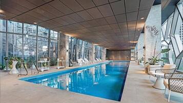 An indoor swimming pool with clear glass walls, lounge chairs, and potted plants.
