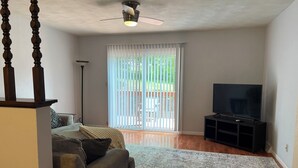 Un salon comprenant un canapé, un téléviseur posé sur un meuble, un ventilateur de plafond et une grande fenêtre avec des stores.