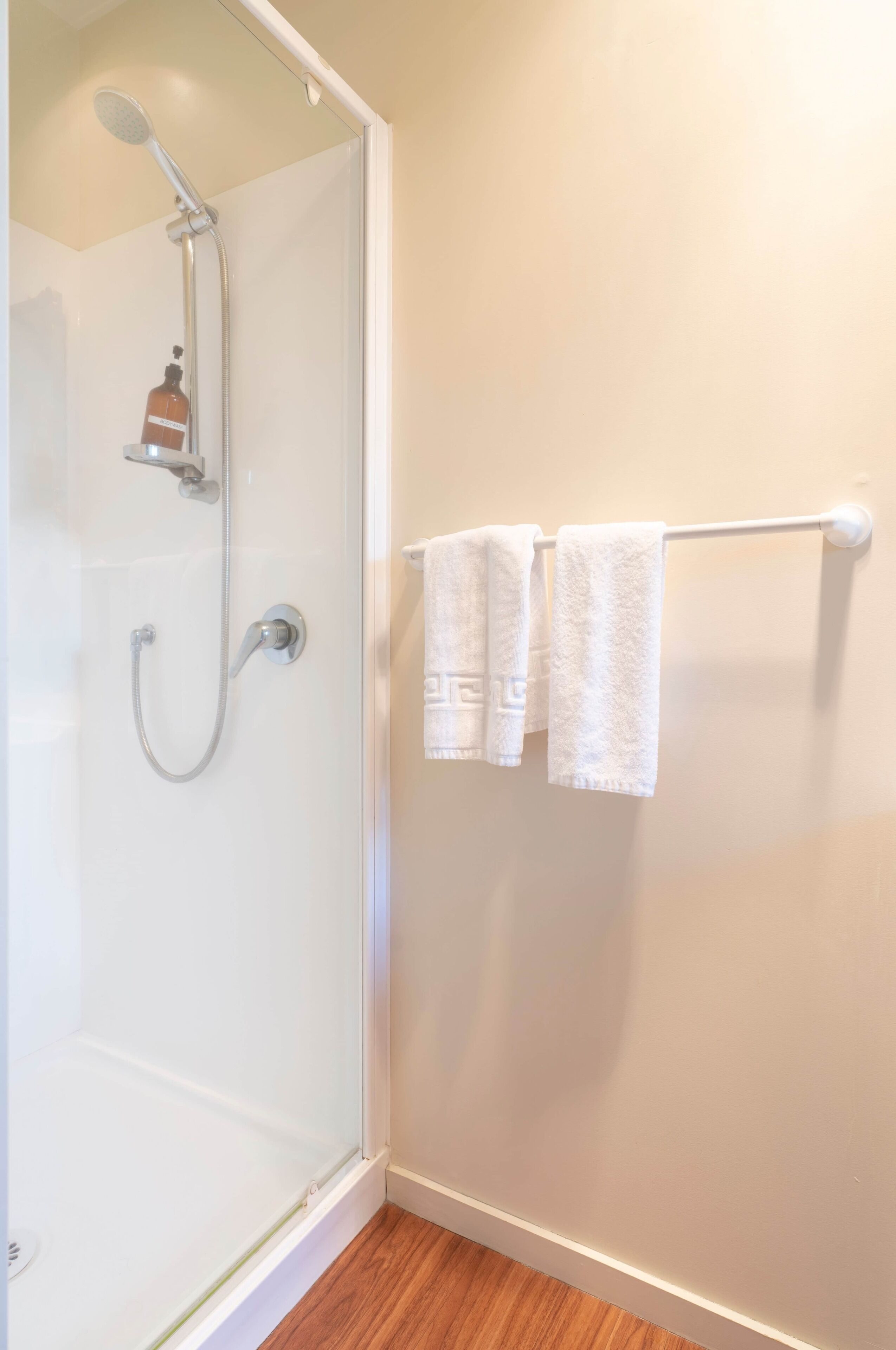 A glass-enclosed shower with a handheld showerhead and a wall-mounted faucet. There are two white towels hanging on a towel rack.