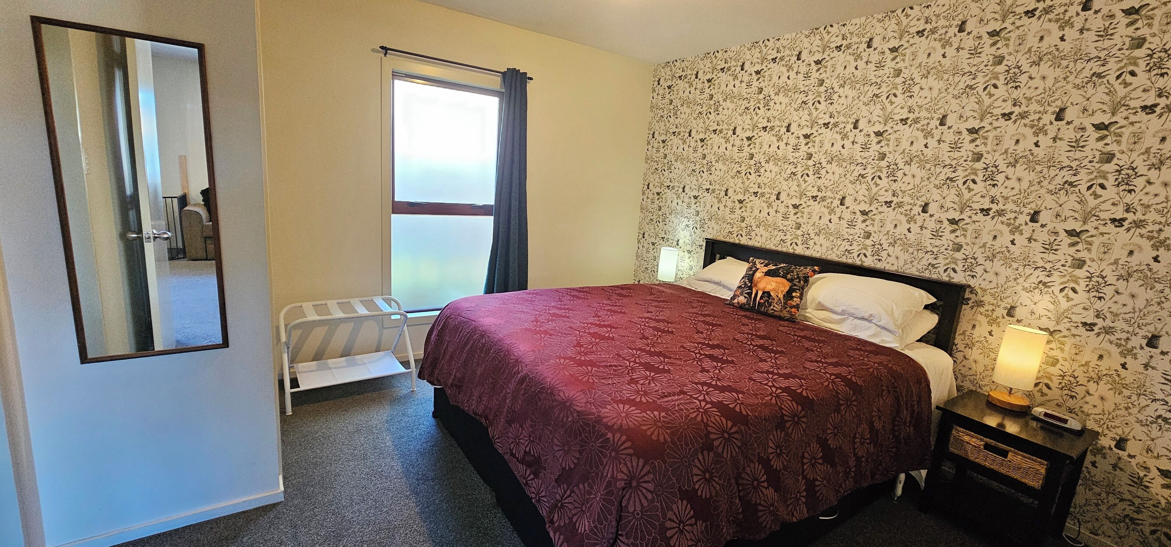 A bedroom with a bed, a nightstand, a lamp, a radiator, a window with curtains, and a floral wallpaper.