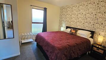 A bedroom with a bed, a nightstand, a lamp, a radiator, a window with curtains, and a floral wallpaper.