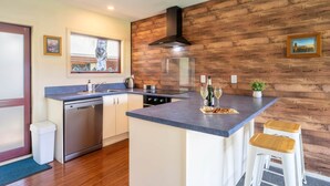 A modern kitchen with a wooden accent wall, a built-in dishwasher, a large island with bar stools, and a window with a view of trees. - Cozy | Functional | Private - HS65214 (Hanmer Springs)