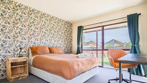 A bedroom with a large bed, a wooden nightstand, an orange chair, and a view of houses through a large window. - Cozy | Functional | Private - HS65214 (Hanmer Springs)
