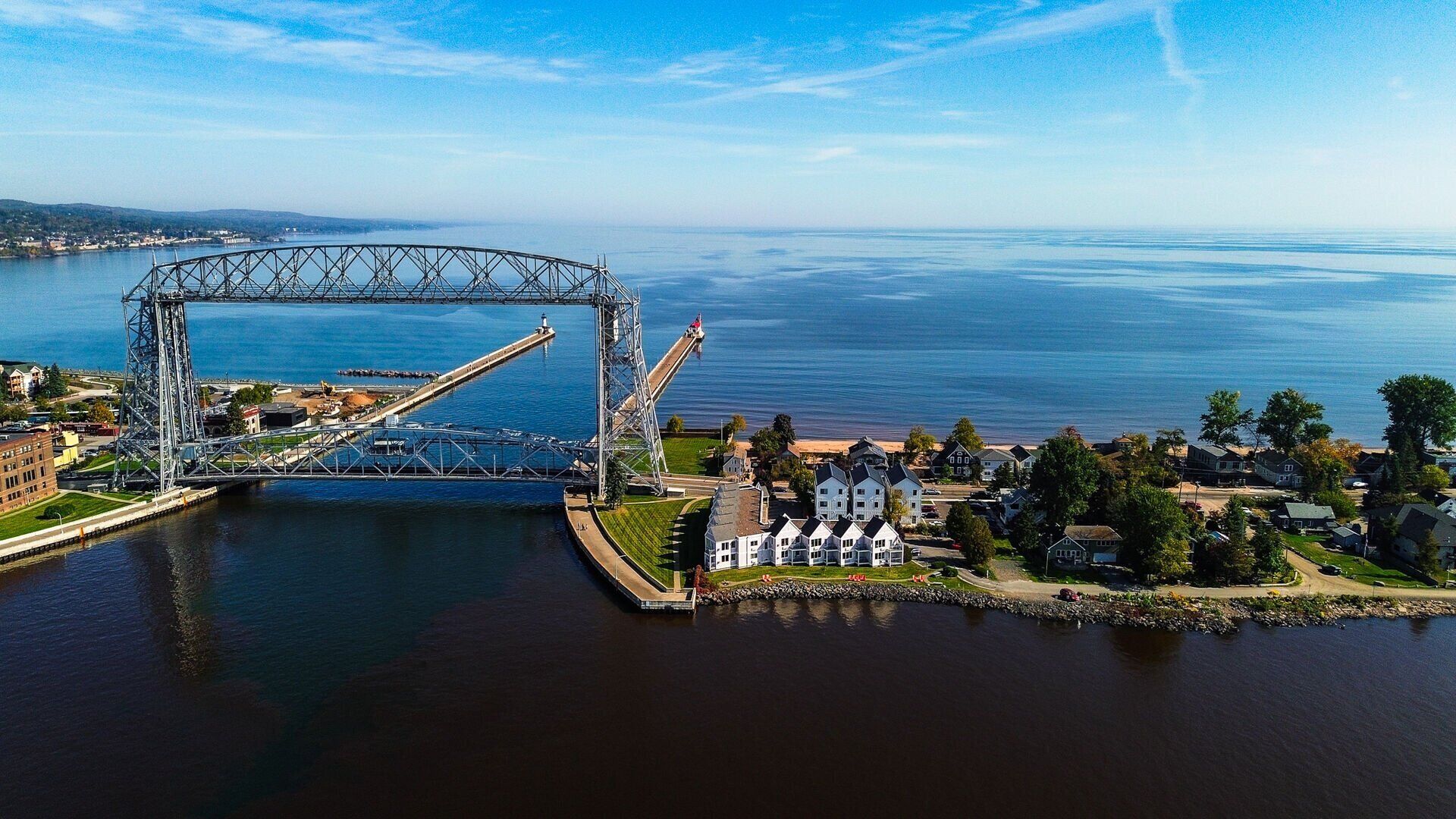 A large steel bridge over a river with a residential area and a lighthouse on the right.