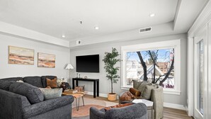 A modern living room with a grey sofa set, a coffee table, and a TV mounted on the wall. There is a large window with a view of buildings outside. - Luxury Condo w/ Beach Access | Dragestil 715-2 (Duluth)